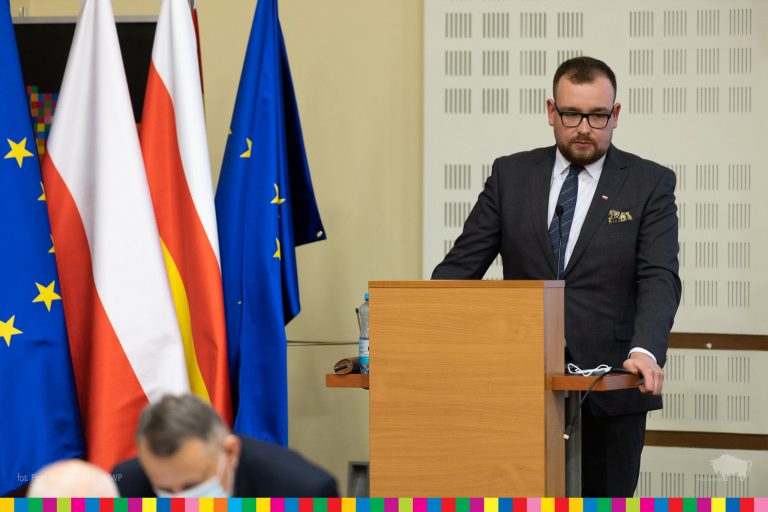 Przy mówinicy stoi mężczyzna w okularach leczniczych i ciemnym garniturze. Z lewej strony widoczna jest flaga Unii Europejskiej., Polski i województwa podlaskiego