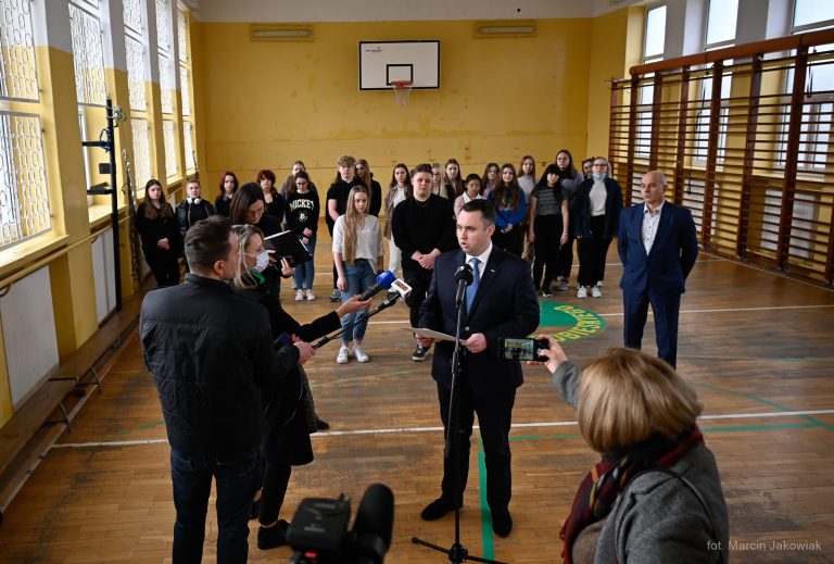 W sali gimnastycznej stoją uczniowie, dyrektor szkoły i prezydent, który mówi do mikrofonów dziennikarzy