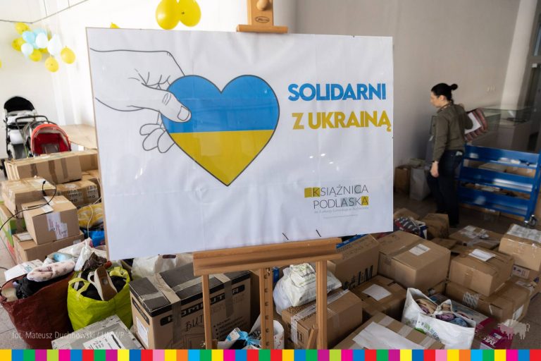 Na drewnianej sztaludze stoi plakat; na białym tle dłoń wystająca z lewej strony trzyma serce, które od góry do połowy figury jest niebieskie, a pod spodem żółte. Obok serca jest niebiesko-żółty napis : Solidarni z Ukrainą. Wokół sztalugi, znajdują się kartony z darami