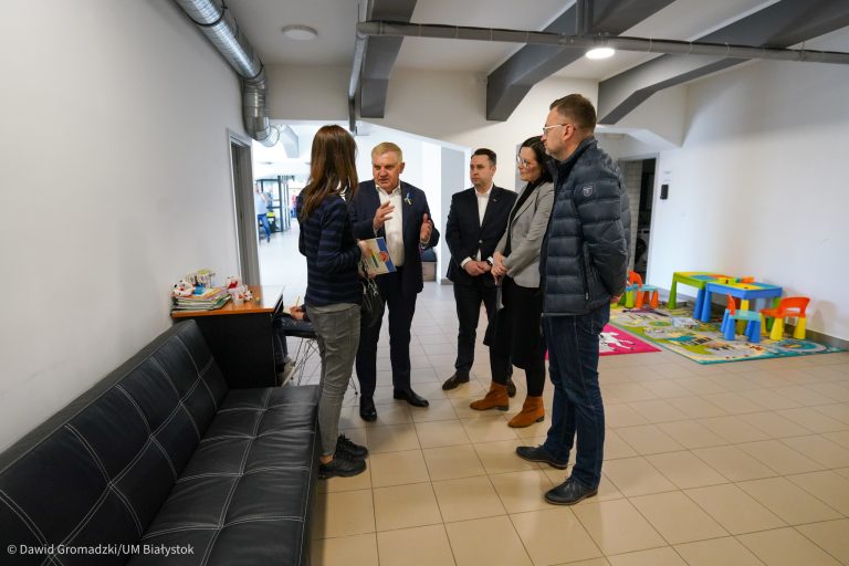 Piec osób stoi w półotwartym kręgu i rozmawiają. Znajdują się w pomieszczeniu industrialnym, z białymi ścianami, pod ścianą z lewej stoi czarna, skórzana kanapa. W tle, w prawym rogu widoczny jest kącik zabaw dla dzieci
