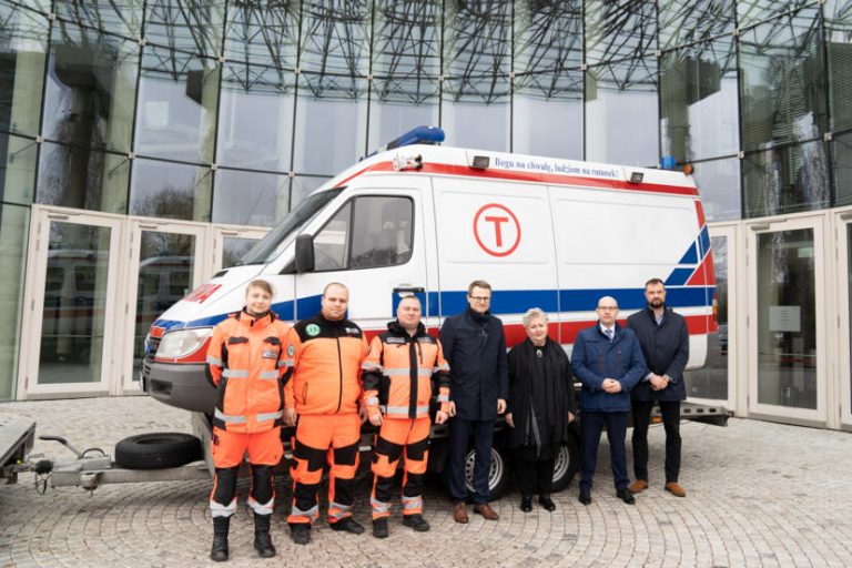 Karetka pogotowia. Przed nią kilkoro osób ustawionych w rzędzie.