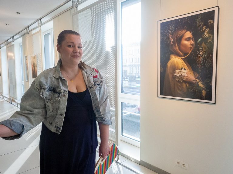 Kobieta w granatowej sukience, dżinsowej kurtce, ze związanymi włosami i podłużną torebką w kolorowe paski w lewej ręce stoi przy jednej ze swoich prac, która wisi na jasnej ścianie