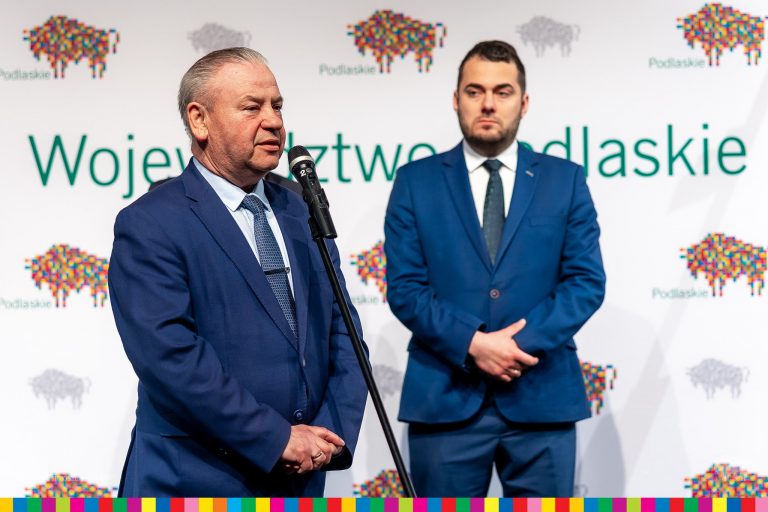 Mężczyzna w granatowym garniturze przemawia do mikrofonu. Stoi on na tle białej ścianki w pikselowe, kolorowe żubry z zielonym napisem: województwo podlaskie. Na tej ściance z prawej strony stoi drugi mężczyzna w garniturze w morskim kolorze