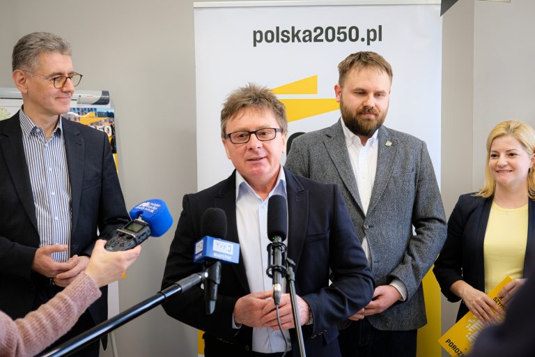 Niewysoki mężczyzna, w okularach mówi do mikrofonu, które przed nim trzymają dziennikarze. Za nim stoi jeszcze dwóch innych mężczyzn, z lewej strony i po środku, a z prawej stoi kobieta