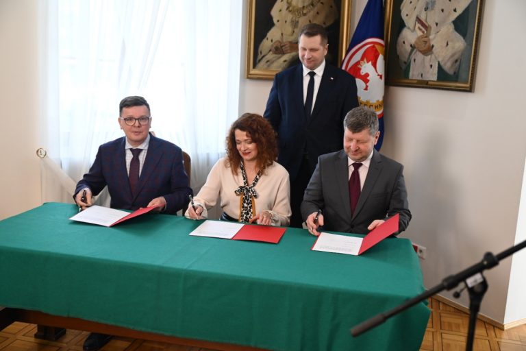 Przy stole zakrytym zielonym suknem siedzą trzy osoby. Po środku kobieta, po dwóch stronach mężczyźni. Pospisują dokumenty. Za kobietą stoi uśmiechnięty mężczyzna, minister edukacji Przemysław Czarnek