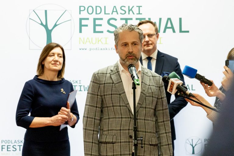 Mężczyzna w ciemno-beżowej marynarce w kratkę stoi przy mikrofonie, do którego mówi. Za nim z lewej strony stoi kobieta, z prawej mężczyzna