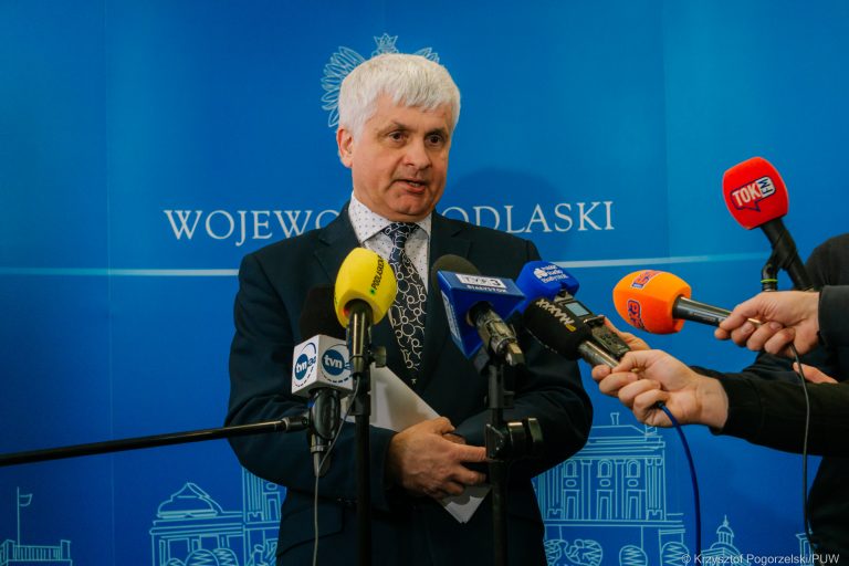 Na niebieskiej ściance z białym napisem: wojewoda podlaski, stoi siwy mężczyzna, w ciemnej marynarce i mówi do mikrofonów, które przed nim trzymają dziennikarze