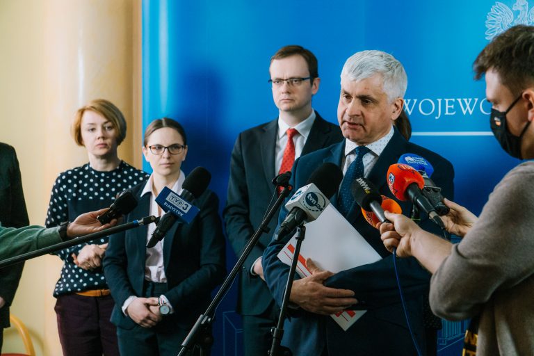 Mężczyzna mówi do mikrofonów , króre na wysokości jego ust trzymają dziennikarze z różnych redakcji . Za nim jest widoczny fragment niebieskiej ścianki. Z jego lewej strony stoją trzy osoby; dwie kobiety, jeden mężczyzna