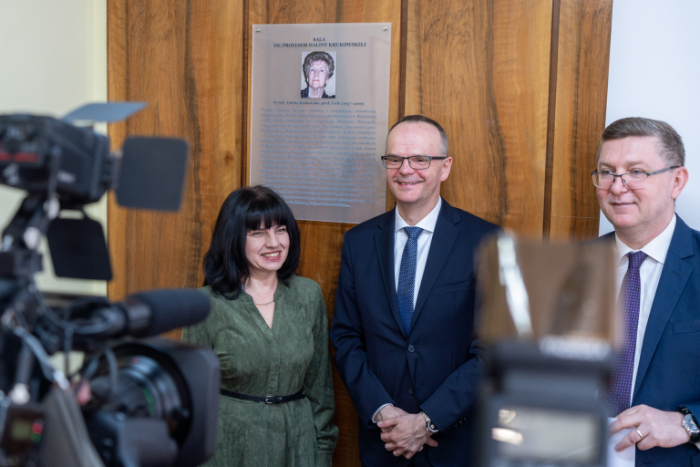 Na drewnianej ścianie wisi tablica upamiętniająca prof. Halinę Krukowską. Przy niej od lewej strony stoi uśmiechnięta kobieta, po środku uśmiechnięty mężczyzna w granatowej marynarce, z prawej również mężczyzna w okularach i granatowej marynarce