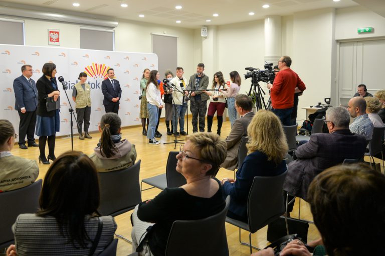 Ludzie mówią do mikrofony podczas konferencji prasowej, przed nimi siedzą osoby na krzesłach