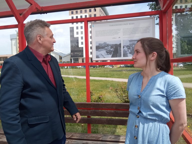 Kobieta i mężczyzna stoją przy tablicy na przystanku autobusowym