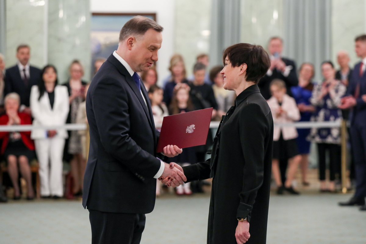Kobieta ubrana na czarno odbiera nominację profesorską od prezydenta Polski, który stoi z lewej strony. Prezydent prawą dłonią ściska dłoń kobiety, w lewej trzyma bordową ze złotym orłem teczkę