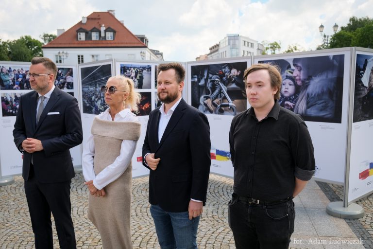 Na tle zdjęć na wystawie stoi trzech mężczyzn i kobieta