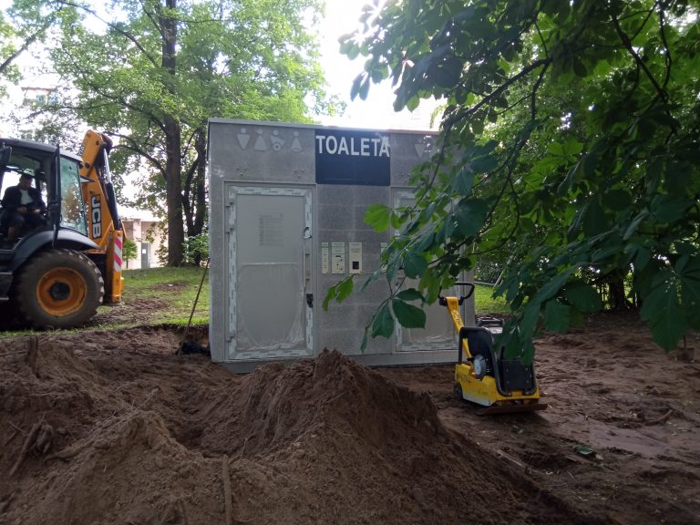 Na ziemi stoi budynek toalety, wokół trwają prace