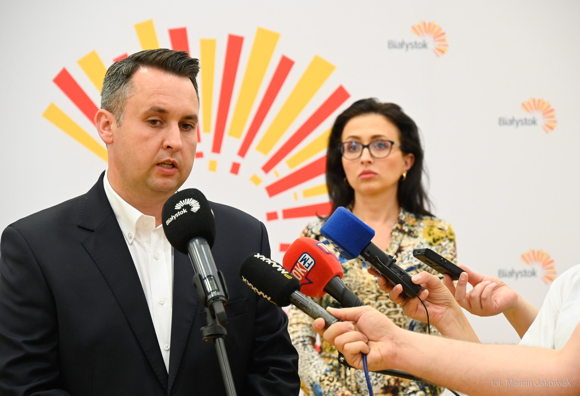 Mężczyzna mówi do mikrofonu, w tle stoi kobieta