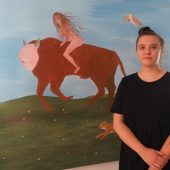 Młoda kobieta stoi przy ścianie, na której wisi jej praca artystyczna