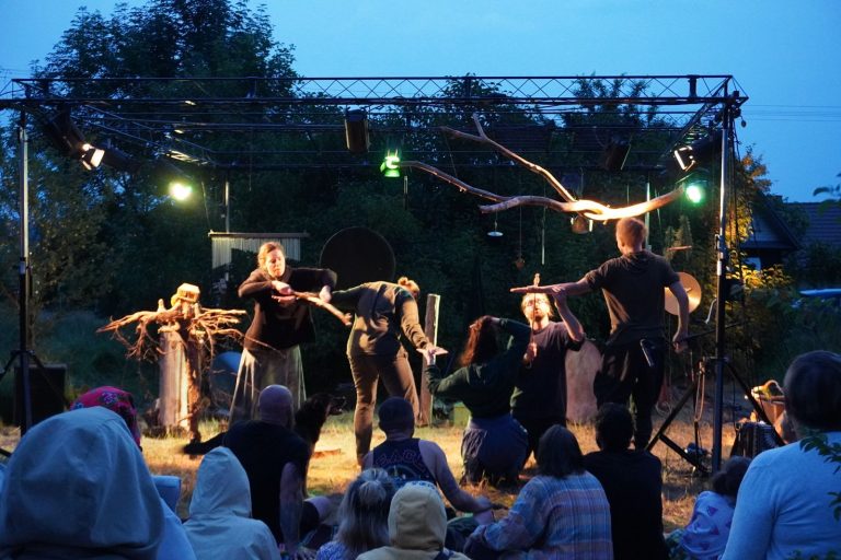 grupa ludzi na scenie pod chmurką o zmroku, przed nimi widownia