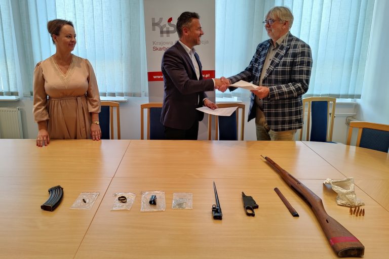Na stole elementy broni. Za stołem kobieta oraz dwóch mężczyzn. Mężczyźni podają sobie dłoń i przekazują dokumenty.