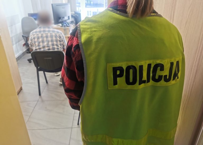 Zatrzymany mężczyzna siedzi na krześle, za nim stoi policjantka w cywilnym ubraniu, w kamizelce z napisem policja.