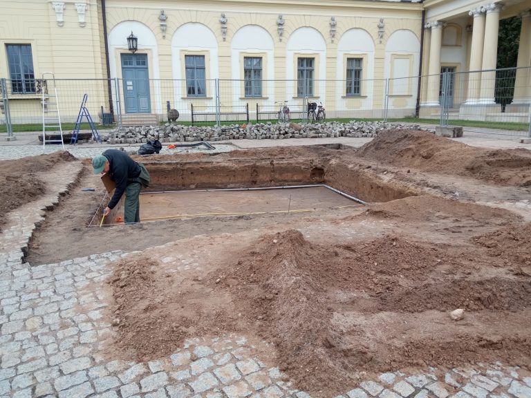 Ogrodzony wykop archeologiczny na parkingu, w wykopie badania prowadzi mężczyzna