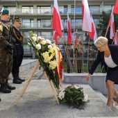 Kobieta elegancko ubrana odkłada kwiaty pod pomnikiem, za nią widać flagi Polski, po lewej stronie stoją żołnierze