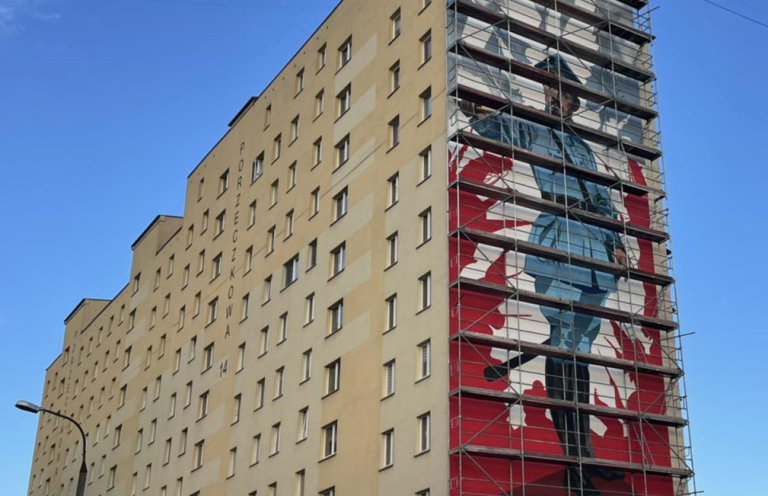 Wierzowiec, na którego szczycie jest namalowany mural z postacią generała, jest ustawione przy muralu rusztowanie.