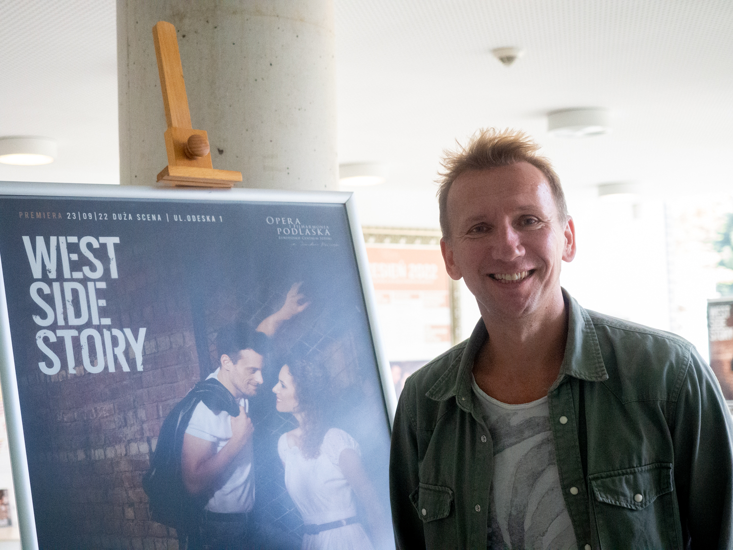 Uśmiechnięty mężczyzna stoi obok plakatu na sztaludze, na plakacie jest tytuł West Side Story i zdjęcie pary patrzącej sobie w oczy