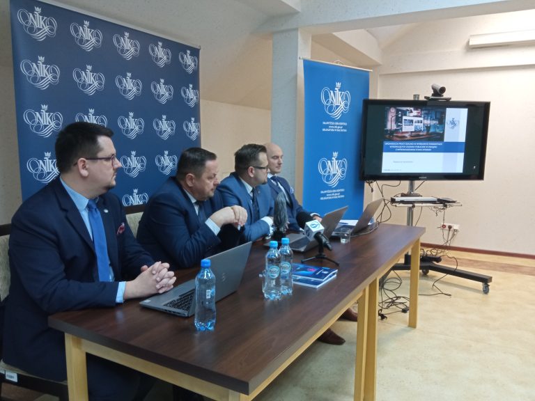 Czterech mężczyzn siedzi za stołem z laptopami i mikrofonami, w tle slajdy na telebimie