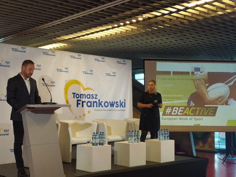Mężczyzna stoi za mównicą przed mikrofonem, obok stoi kobieta