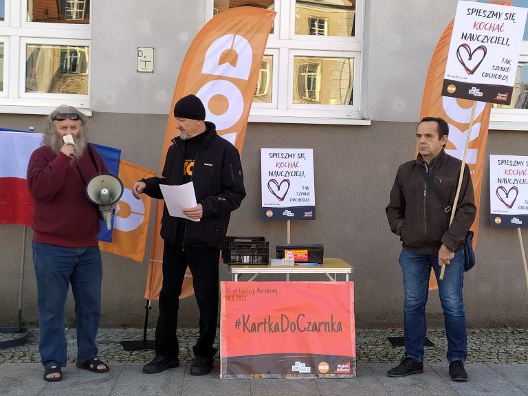 Trzech mężczyzn stoi pod budynkiem, jeden z nich trzyma megafon, drugi transparent. Z tyłu widać flagi.