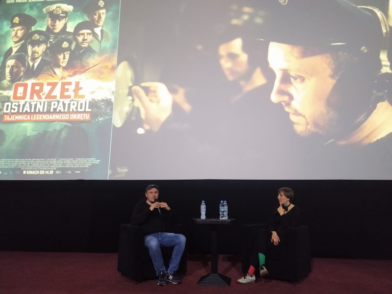 Kobieta i mężczyzna siedzą przy małym stoliku i trzymają mikrofony, a za nimi na ekranie wyśietla się kadr z filmu