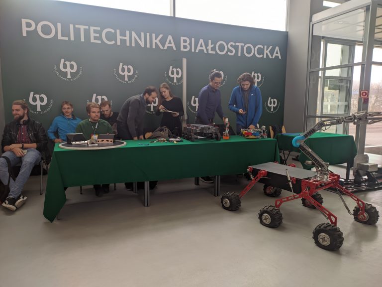 Młodzi ludzie stoją i siedzą za stołem, przed nimi jest łazik marsjański