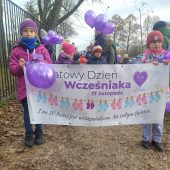 Dziewczynki trzymają transparent z napisem Światowy Dzień Wcześniaka, za nimi dzieci trzymają fioletowe balony