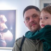 Mężczyzna trzyma na rękach synka kilkuletniego, w tle wystawa fotografii