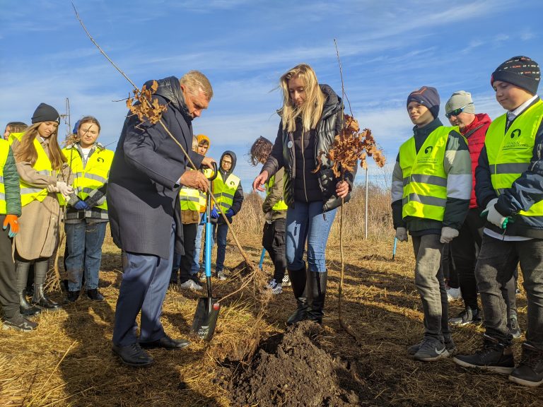 Kobieta i mężczyzna sadzą drzewa, dookoła nich stoją dzieci w odblaskowych kamizelkach.