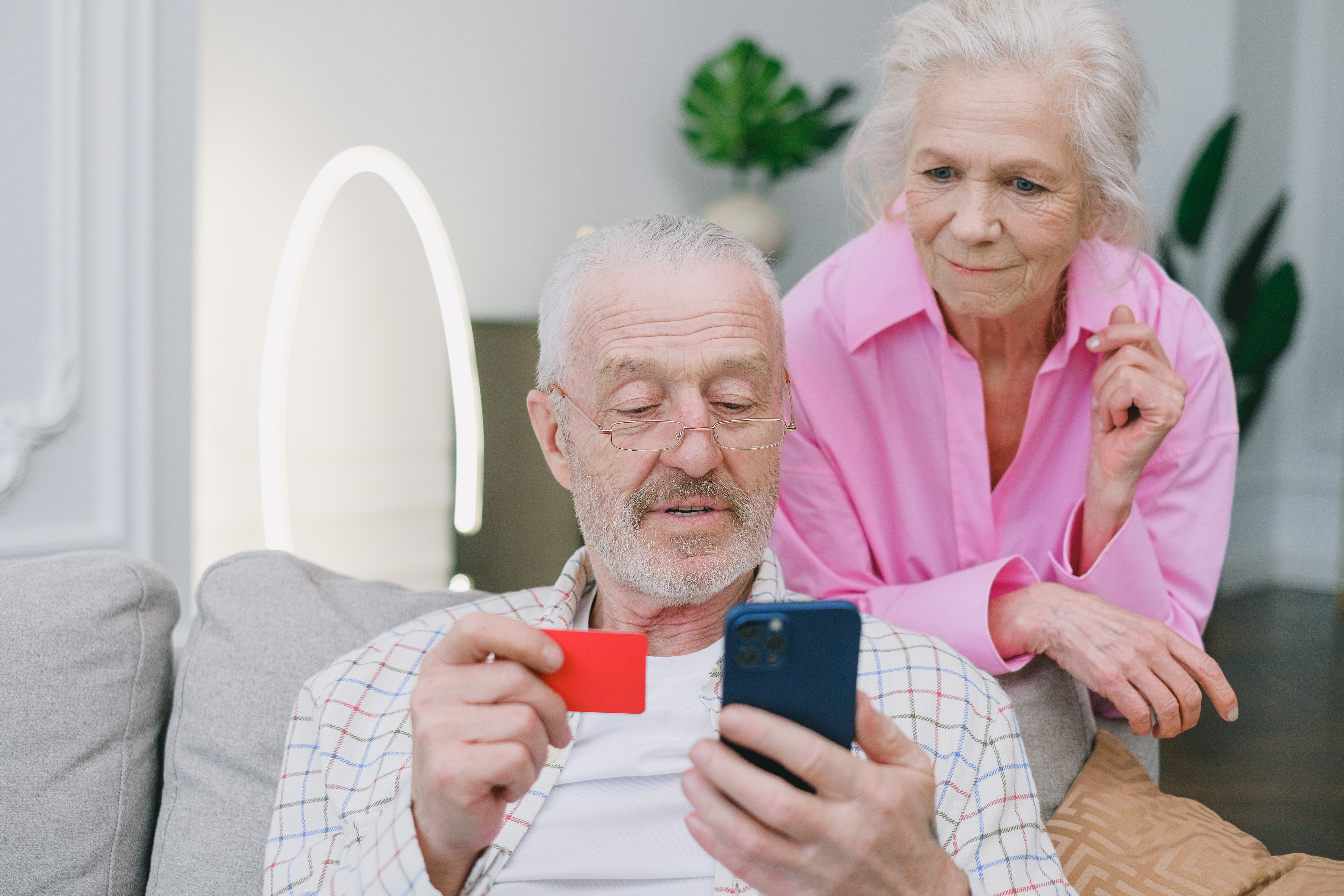 Starszy męźczyzna trzyma kartę płatniczą i telefon komórkowy, starsza kobieta to obserwuje
