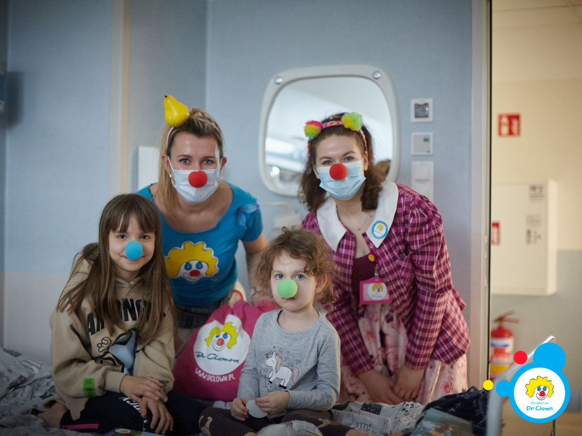 Dwie kobiety w kolorowych strojach pozują do zdjęcia z dwójką dzieci w szpitalnej sali. Wszyscy mają kolorowe piłeczki na nosach.