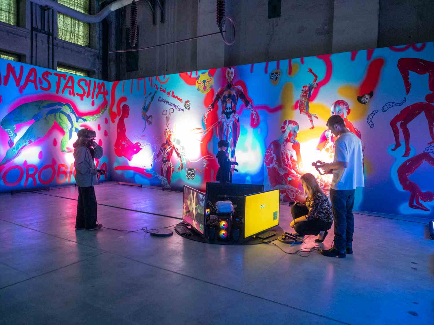 Wystawa z kolorowymi podświetlonymi pracami wizualnymi, na środku stoją ludzie z okularami do wirtualnej rzeczywistości.