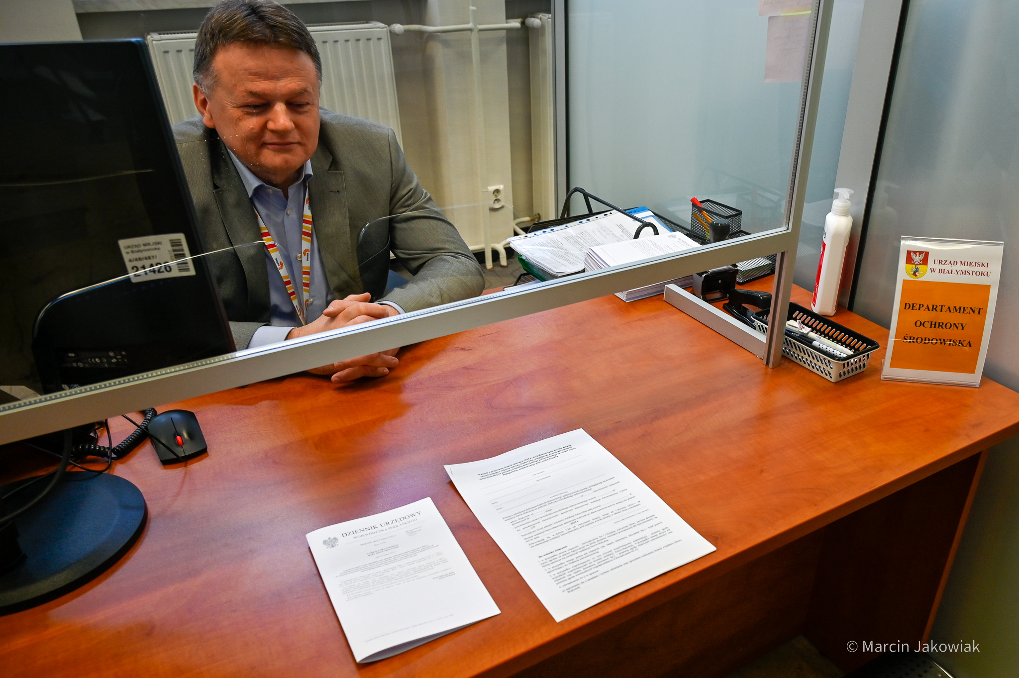 Urzędnik w departamencie ochrony środowiska siedzi w okienku; na biurku leżą druki urzędowe