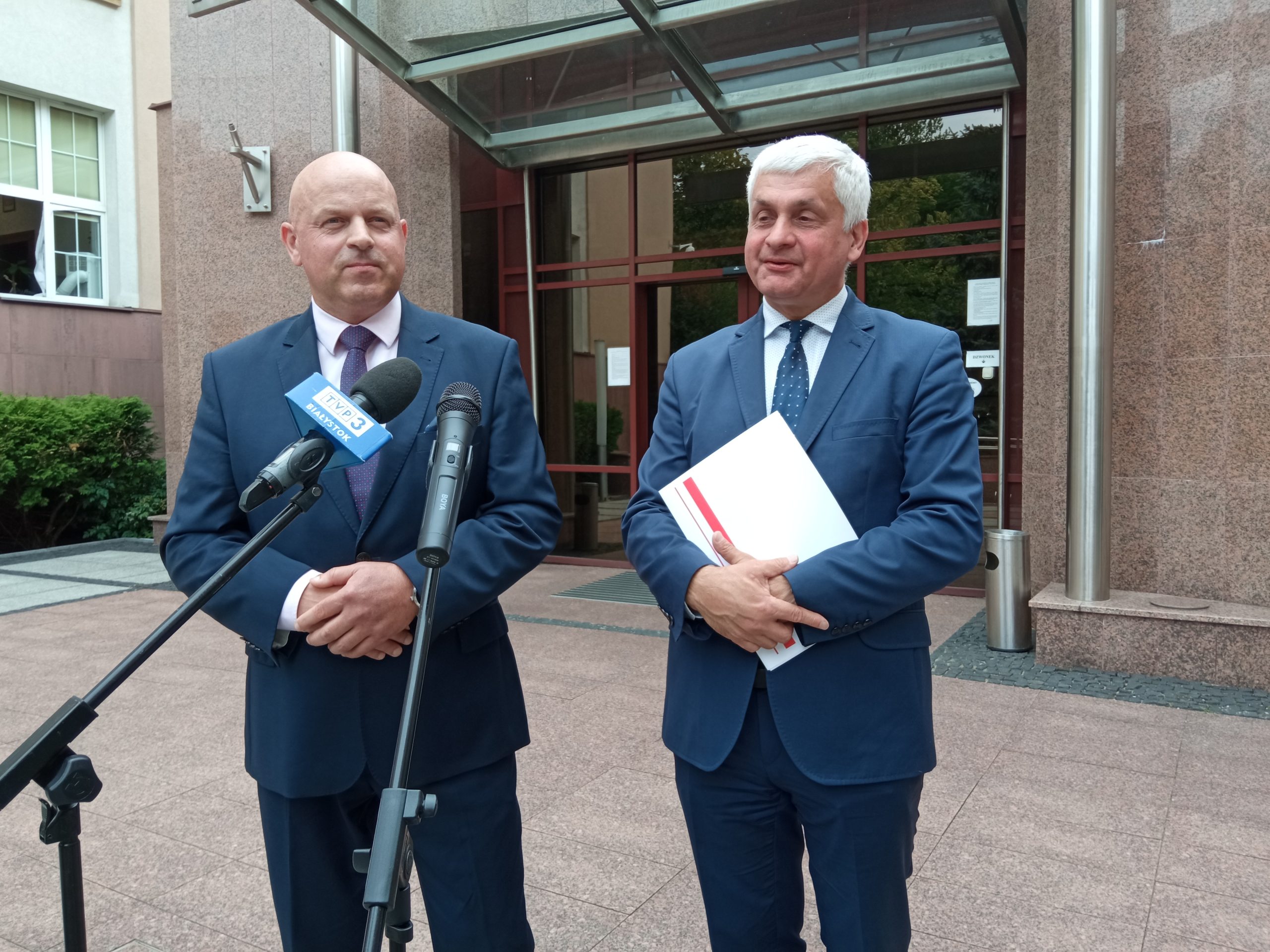 Dwaj mężczyźni stoją przed mikrofonami przed budynkiem