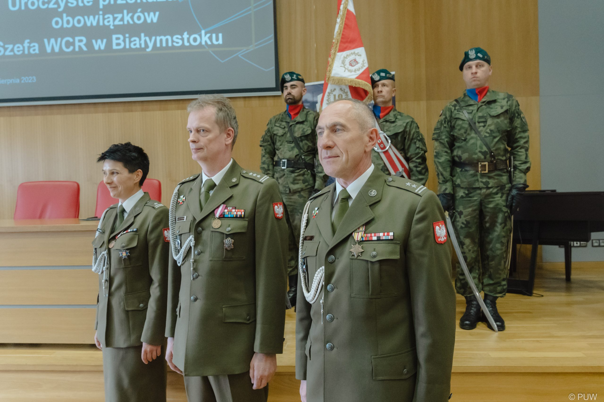 Kobieta i dwóch mężczyzn stoi w wojskowych mundurach, za nimi poczet sztandarowy