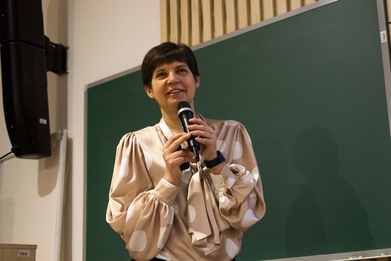 Kobieta o krótkich ciemnych włosach stoi z mikrofonem w dłoni na tle tablicy