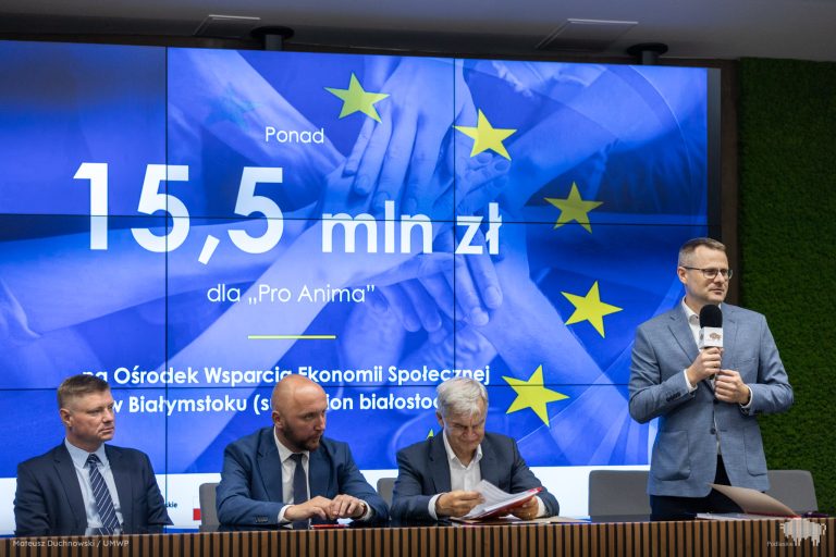 Ludzie siedzą za stołem, a za nimi telebim.