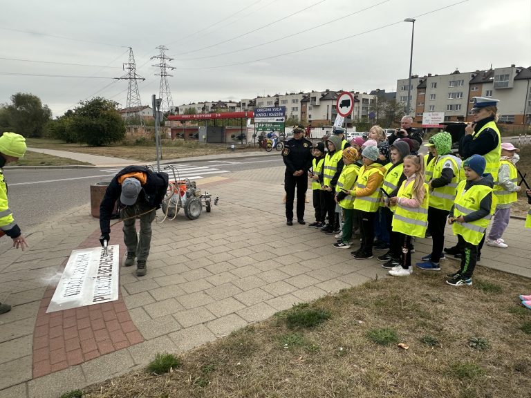 Dzieci w kamizelkach odblaskowych przyglądają się jak panowie malują napis "Odłóż telefon, przejdź bezpiecznie"
