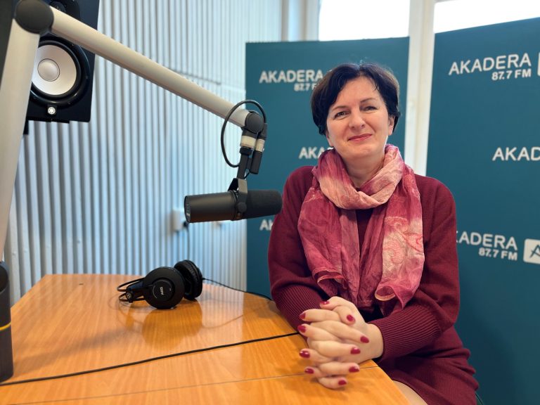 Kobieta w sile wieku o krótkich ciemnych włosach siedzi przy stole, ma szal i czerwony sweter