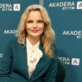 Prof. Katarzyna Halicka podczas wizyty w Radiu AKADERA, siedzi na tle niebieskich plansz z logo stacji, uśmiechnięta, w zielonej marynarce i białej bluzce z kokardą
