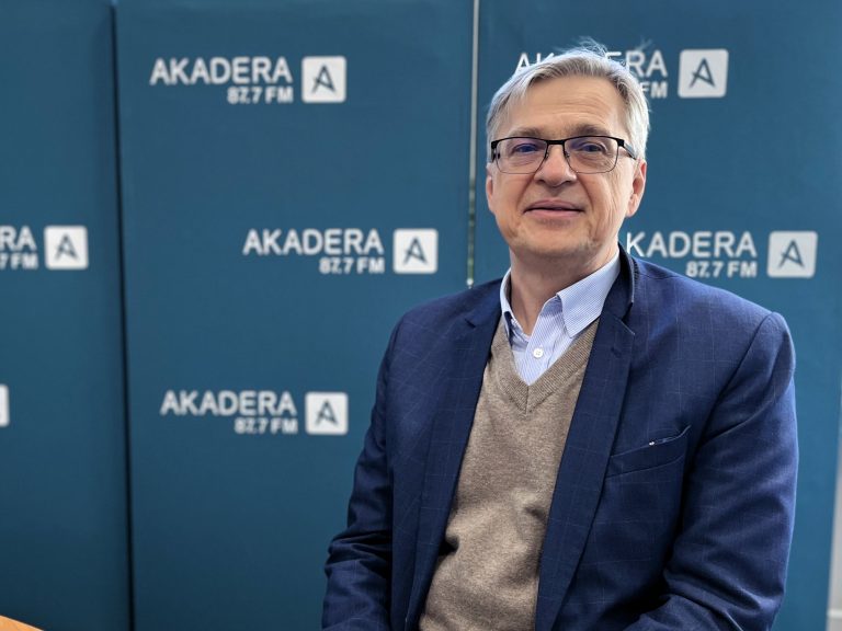 Mężczyzna o siwych włosach w okularach w ciemnej marynarce i jasnym swetrze, lekko uśmiechnięty