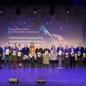 Grupa osób stojących na scenie podczas gali wręczenia stypendiów w dziedzinie twórczości artystycznej, upowszechniania kultury oraz opieki nad zabytkami. W tle duży ekran z grafiką przedstawiającą pędzel malarski oraz tekst informujący o wydarzeniu. Część laureatów trzyma w rękach dyplomy i kwiaty.