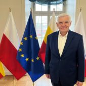 Prezydent Białegostoku Tadeusz Truskolaski stoi na tle flag Polski, Unii Europejskiej oraz województwa podlaskiego. Ma na sobie granatową marynarkę i białą koszulę, uśmiecha się do obiektywu.