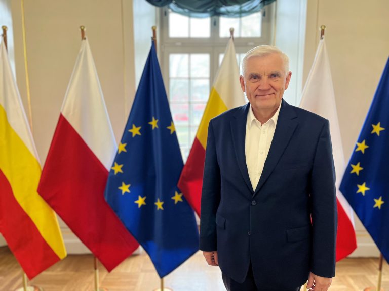 Prezydent Białegostoku Tadeusz Truskolaski stoi na tle flag Polski, Unii Europejskiej oraz województwa podlaskiego. Ma na sobie granatową marynarkę i białą koszulę, uśmiecha się do obiektywu.
